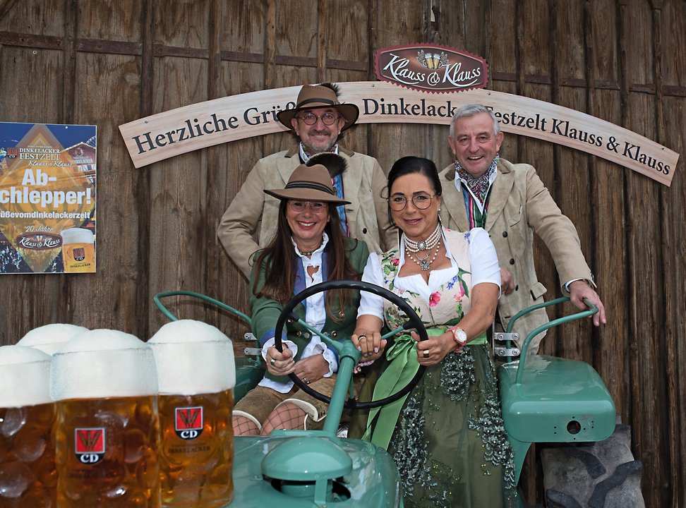 Die Brüder Dieter und Werner Klauss sitzen gemeinsam mit ihren beiden Frauen auf einem grünen Traktor und lächeln in die Kamera. Vorne auf der Motorhaube sind vier Maß Bier von Dinkelacker platziert.