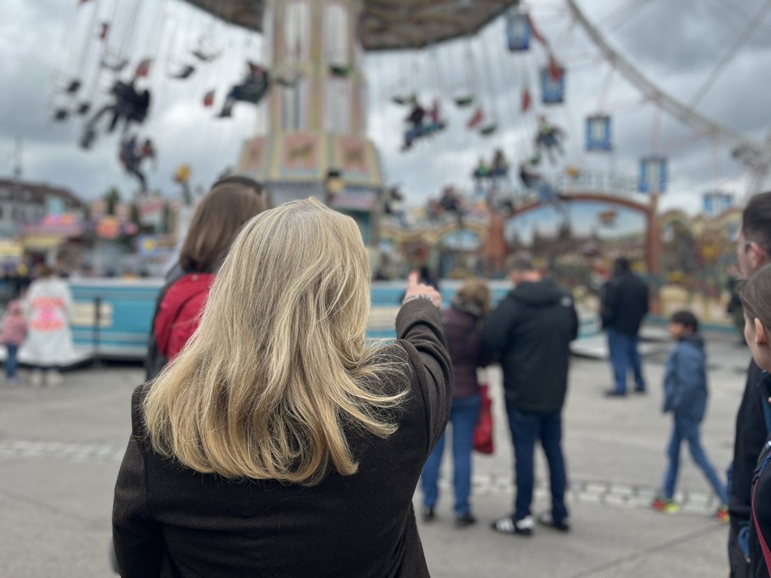 Eine blonde Frau in Tracht führt über den Wasen und zeigt auf etwas an einem Kettenkarussell