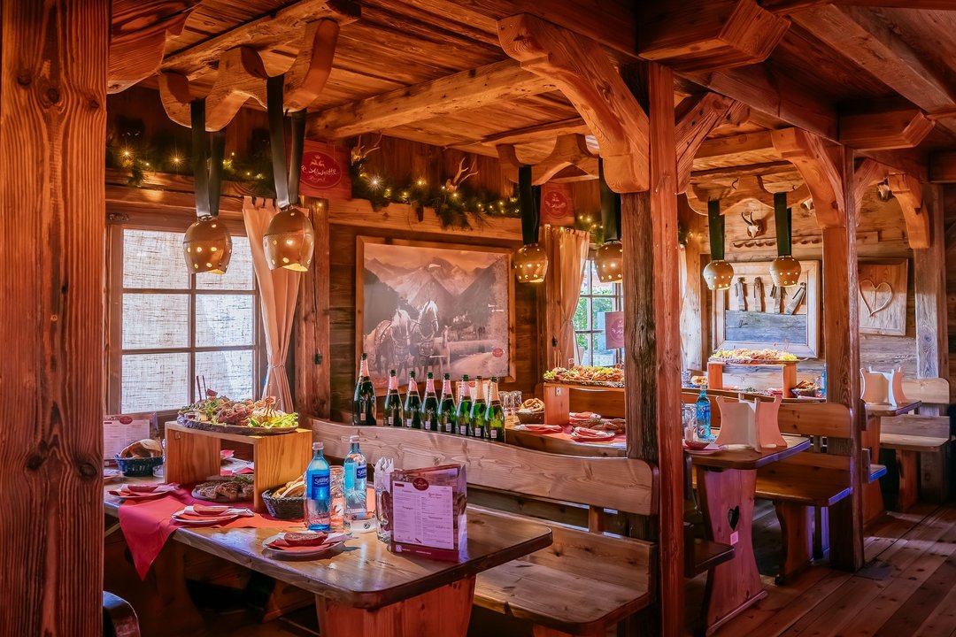 Sitzplätze der Almhütte Royal mit Kuhglicken, die als Lampen von den Decken hängen. Auf den Tischen sind Brotzeit-Bretter schön drapiert. 