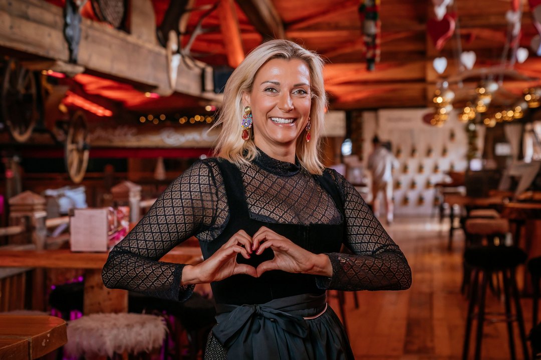 Geschäftsführerin Nina Renoldi im Dirndl in der Almhütte Royal, lächelt in die Kamera und formt mit ihren Händen ein Herz.