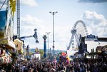 Blick über Feststraße auf Fruchtsäule, Riesenrad, Fahrgeschäfte und viele Menschen.