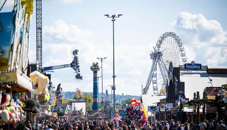 Blick über Feststraße auf Fruchtsäule, Riesenrad, Fahrgeschäfte und viele Menschen.