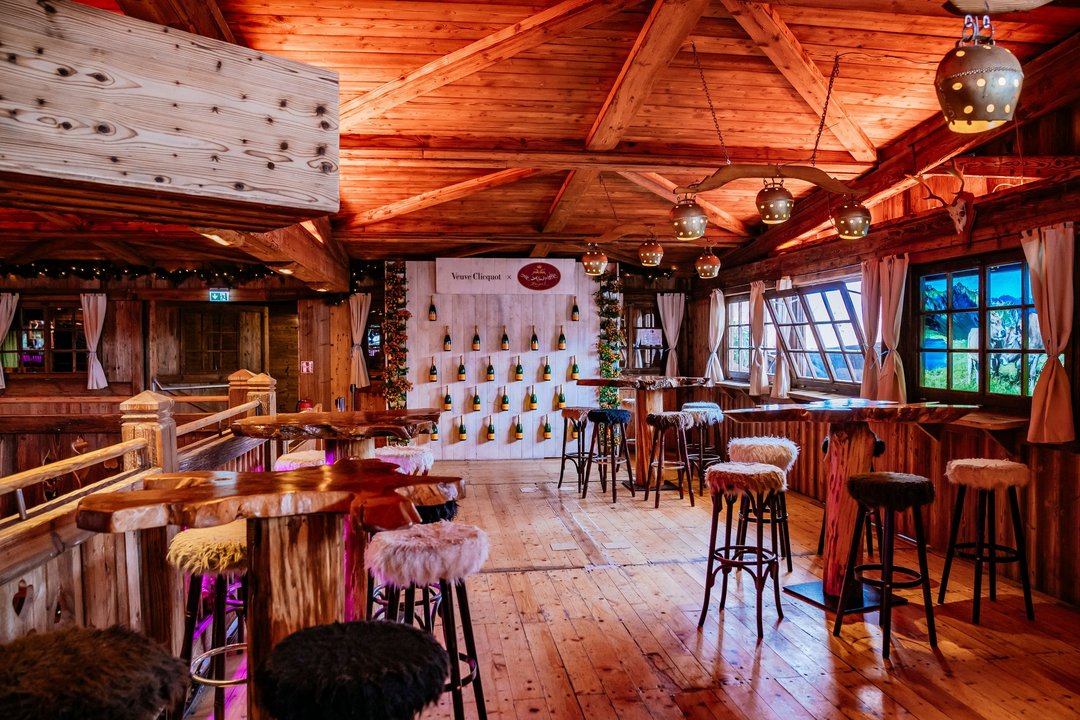 Der Bar-Bereich der Almhütte Royal. An einer Wand hängen lauter Sektflaschen. Drumherum sind Stehtische mit Hochstühlen, die mit Fell überzogen sind.